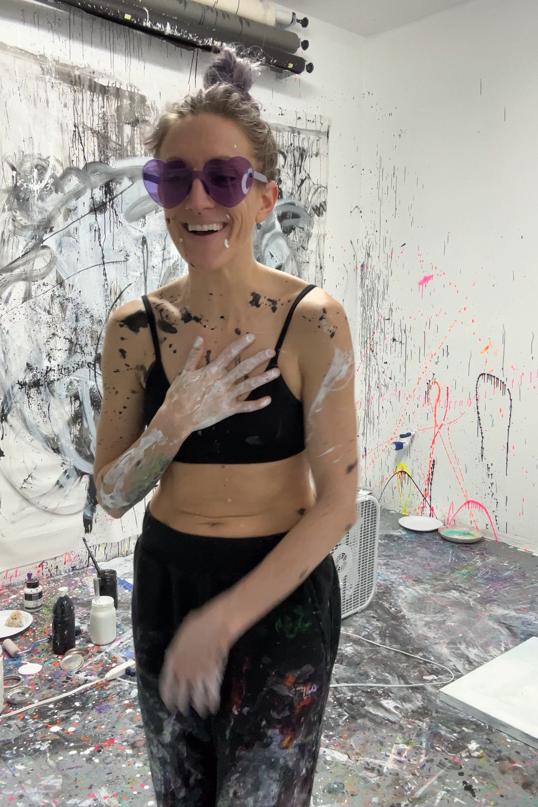 A woman in a paint-splattered studio wearing purple sunglasses, a black sports bra, and black pants, laughing with her hand on her chest. Large abstract canvases and paint-covered walls are visible in the background.
